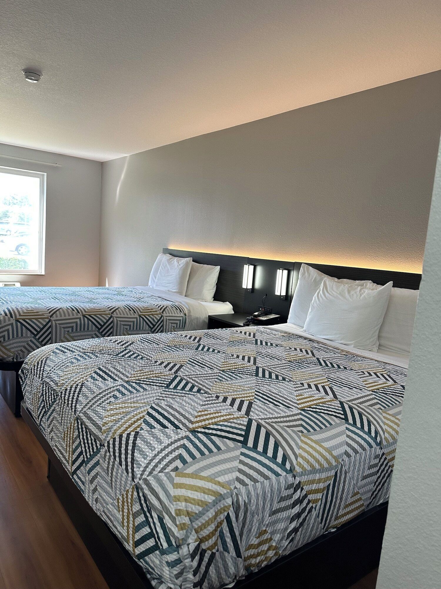A hotel room with two beds, a window, and a headboard with built-in lighting.