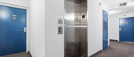 A modern elevator with a metallic finish, a blue door with a number plate, and a wall-mounted emergency phone.
