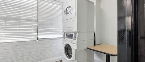 A laundry room with a stacked washer and dryer, a small table, and a window with blinds.