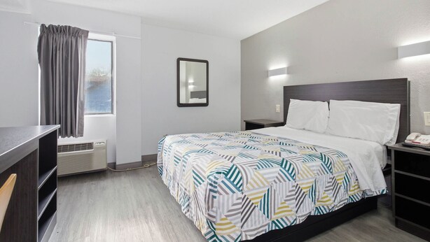 A hotel room with a bed, bedside tables, a desk, a chair, a window with curtains, an air conditioning unit, and a wall-mounted mirror. - Motel 6 Washington, DC (Washington)