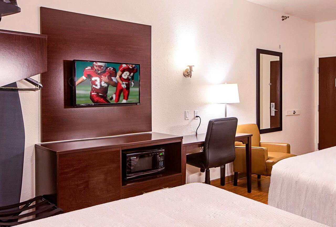 A hotel room with a bed, a desk with a television displaying football players, a microwave, a chair, and a lamp.