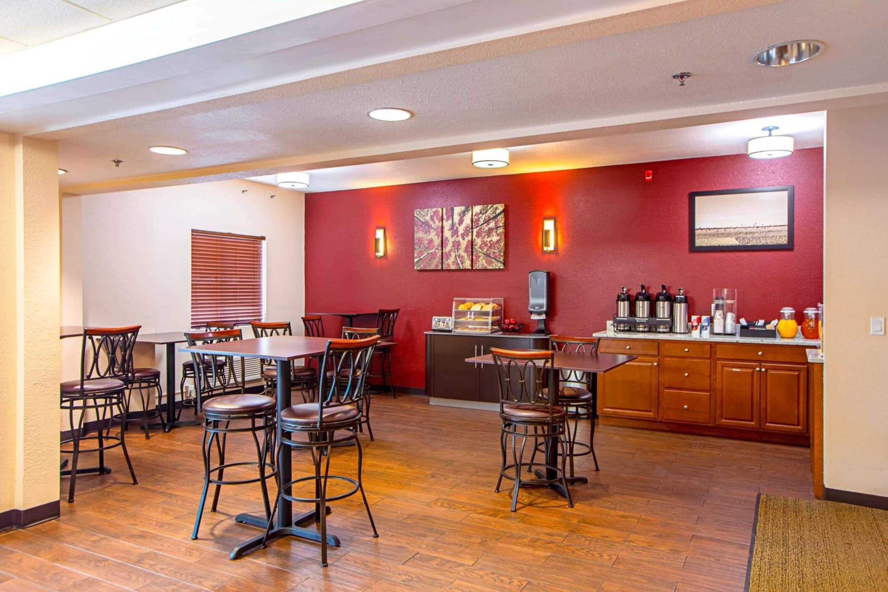 A hotel dining area with wooden tables and chairs, a kitchen counter with appliances, and artwork on the walls.