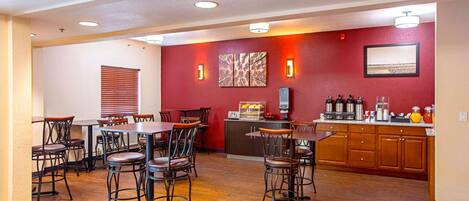 A hotel dining area with wooden tables and chairs, a kitchen counter with appliances, and artwork on the walls.
