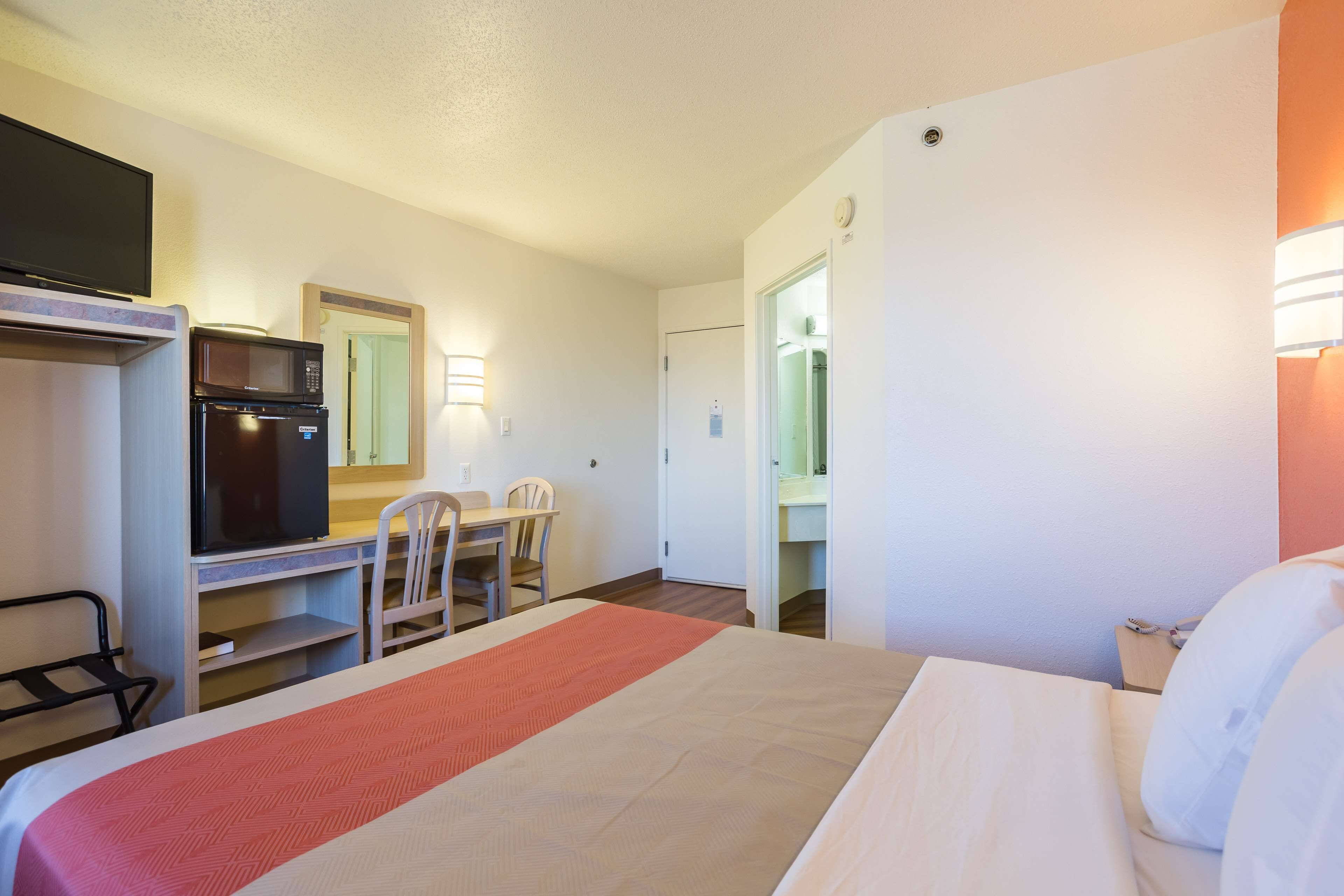 A hotel room with a bed, a desk with a microwave and refrigerator, a TV, and a bathroom visible through an open door.
