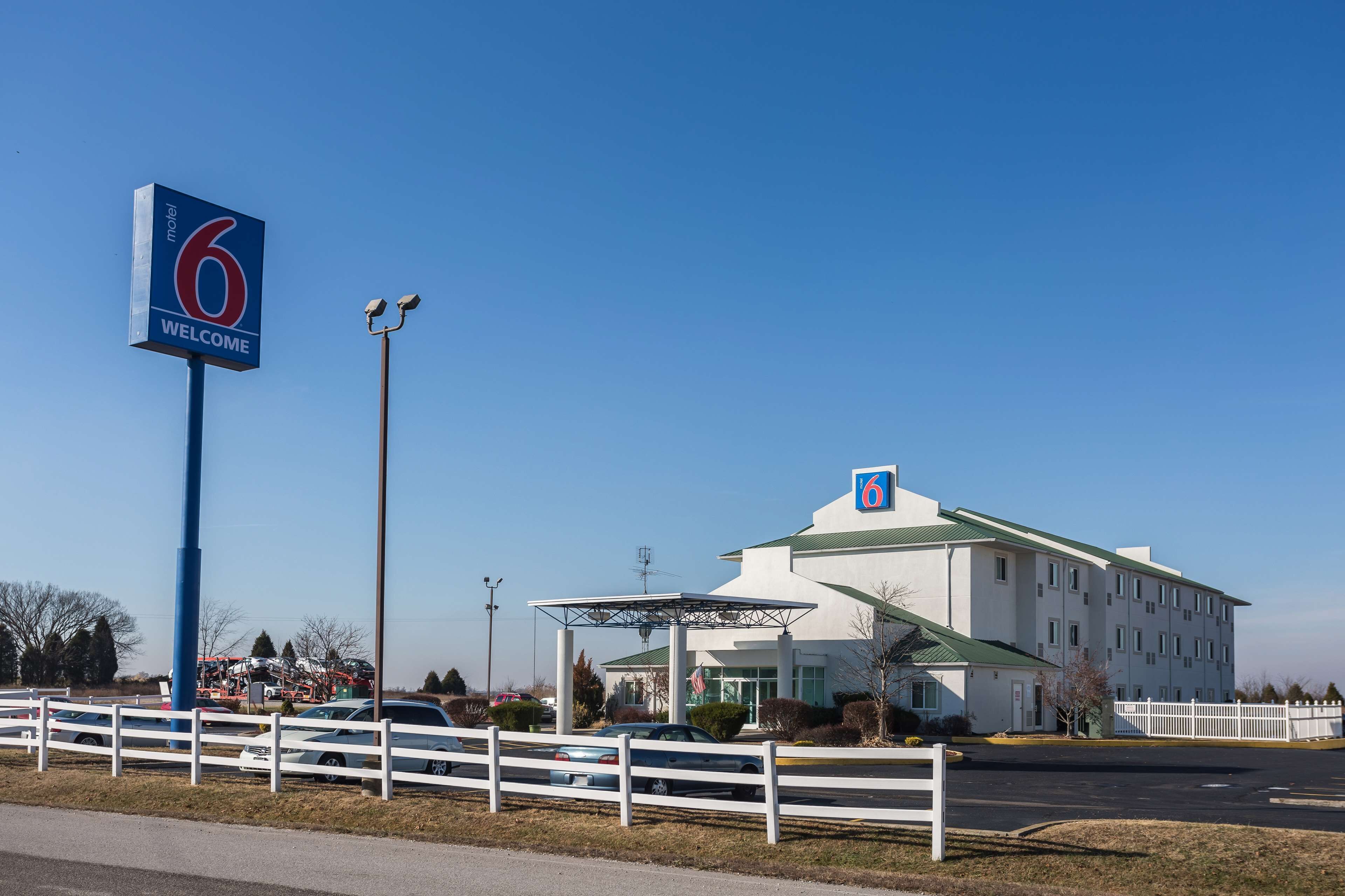 Un motel reconnaissable à son grand panneau indiquant le chiffre « 6 », doté d’un parking couvert et entouré d’une clôture blanche.