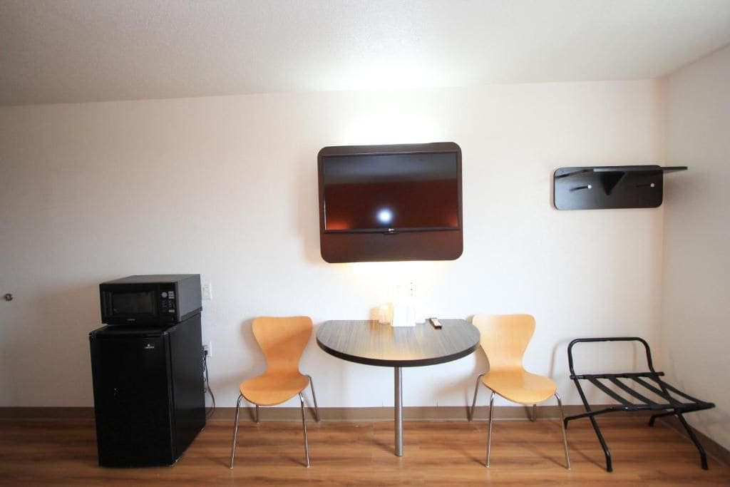 A compact living space with a small kitchenette, a round dining table, two chairs, a flat-screen TV mounted on the wall, and a black shelf.