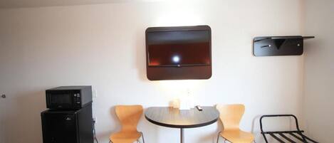 Un espace de vie compact comprenant une petite kitchenette, une table de salle à manger ronde, deux chaises, un téléviseur à écran plat fixé au mur et une étagère noire.