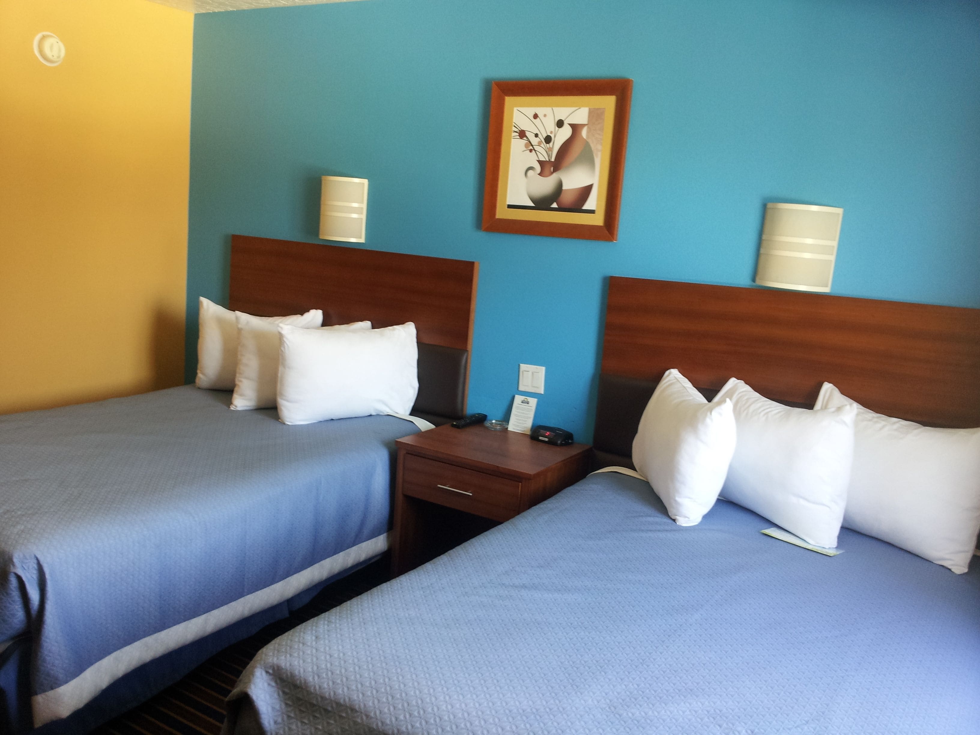 A hotel room with two beds, each with white bedding and a wooden headboard. There is a nightstand between the beds with a lamp and a telephone.