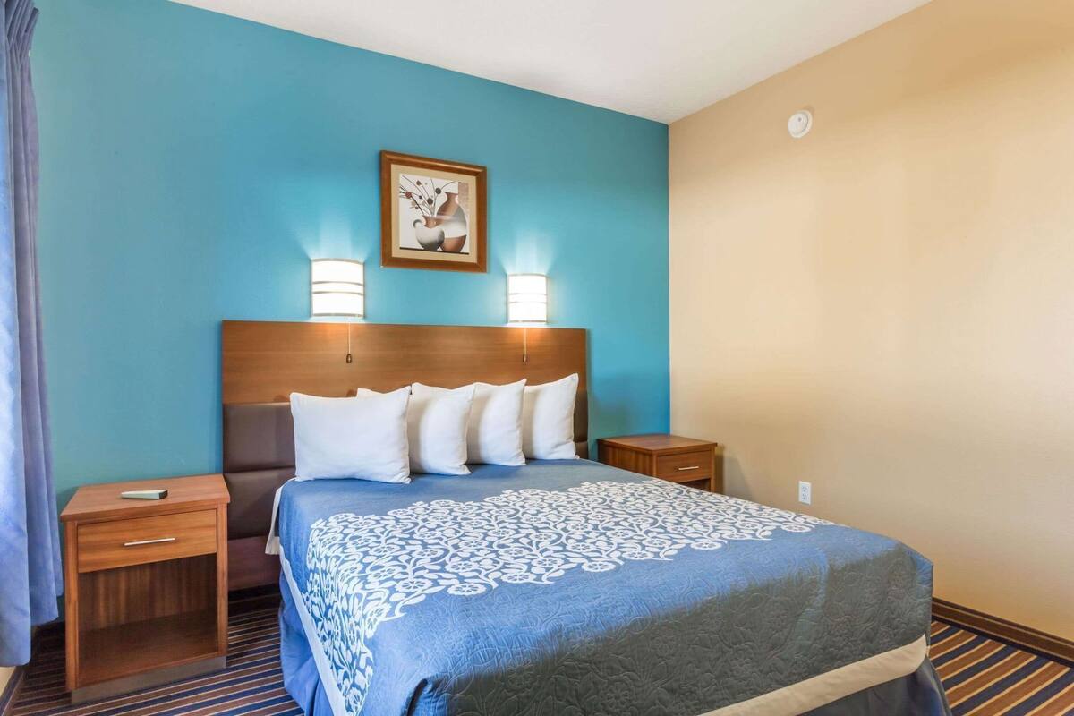 A hotel room with a bed, two bedside tables, a nightstand, a wall-mounted lamp, and a framed picture.