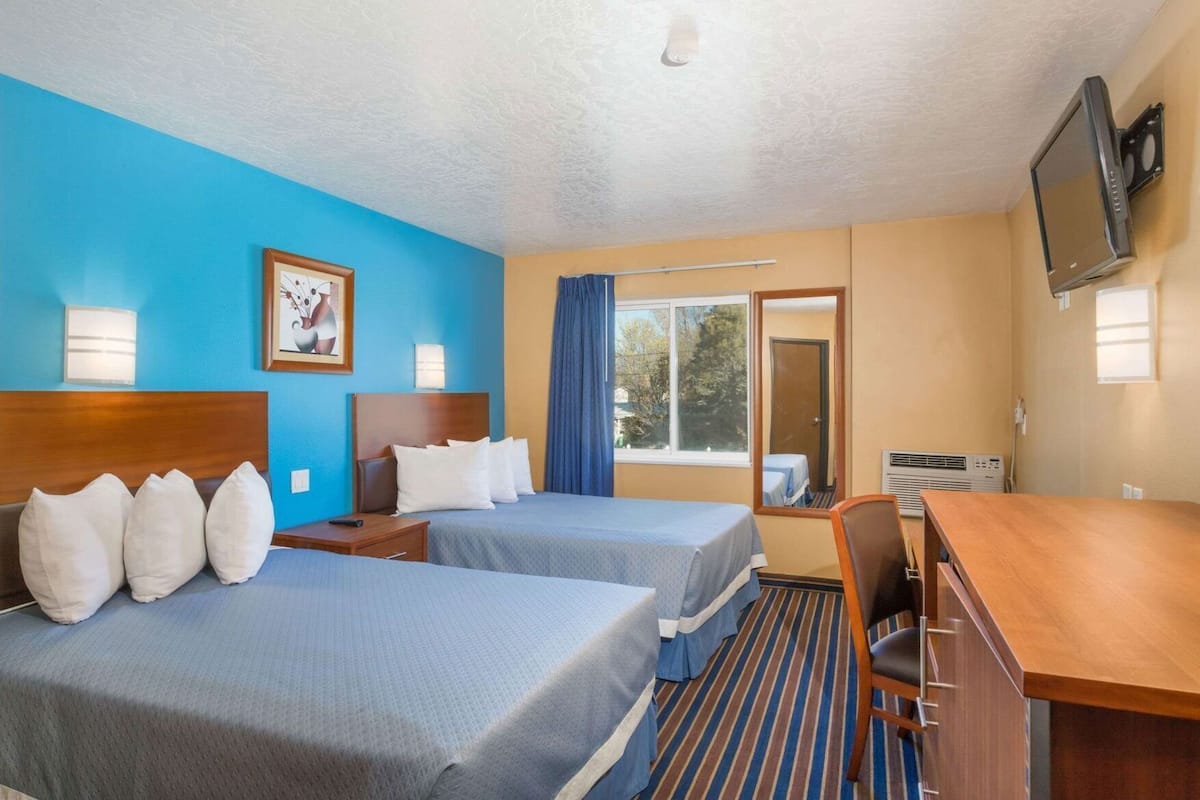 A hotel room with two beds, a desk, a TV, and a window with curtains.
