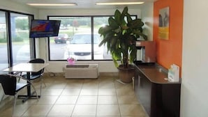 A clean, modern interior with a reception desk, a large potted plant, a flat-screen TV, and a view of the parking lot through large windows. - Motel 6 Columbus, MS (Columbus)
