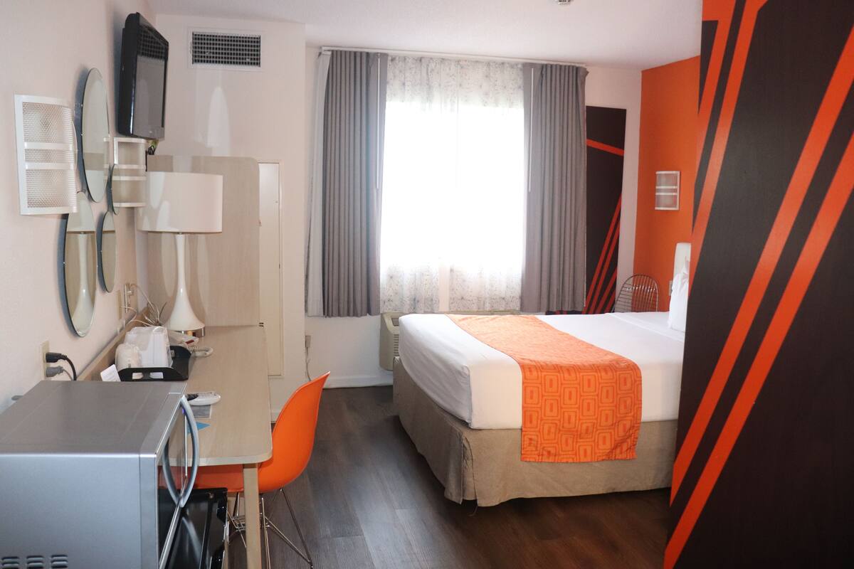 Quarto de hotel com cama, escrivaninha com cadeira, micro-ondas, espelho e janela com cortinas.