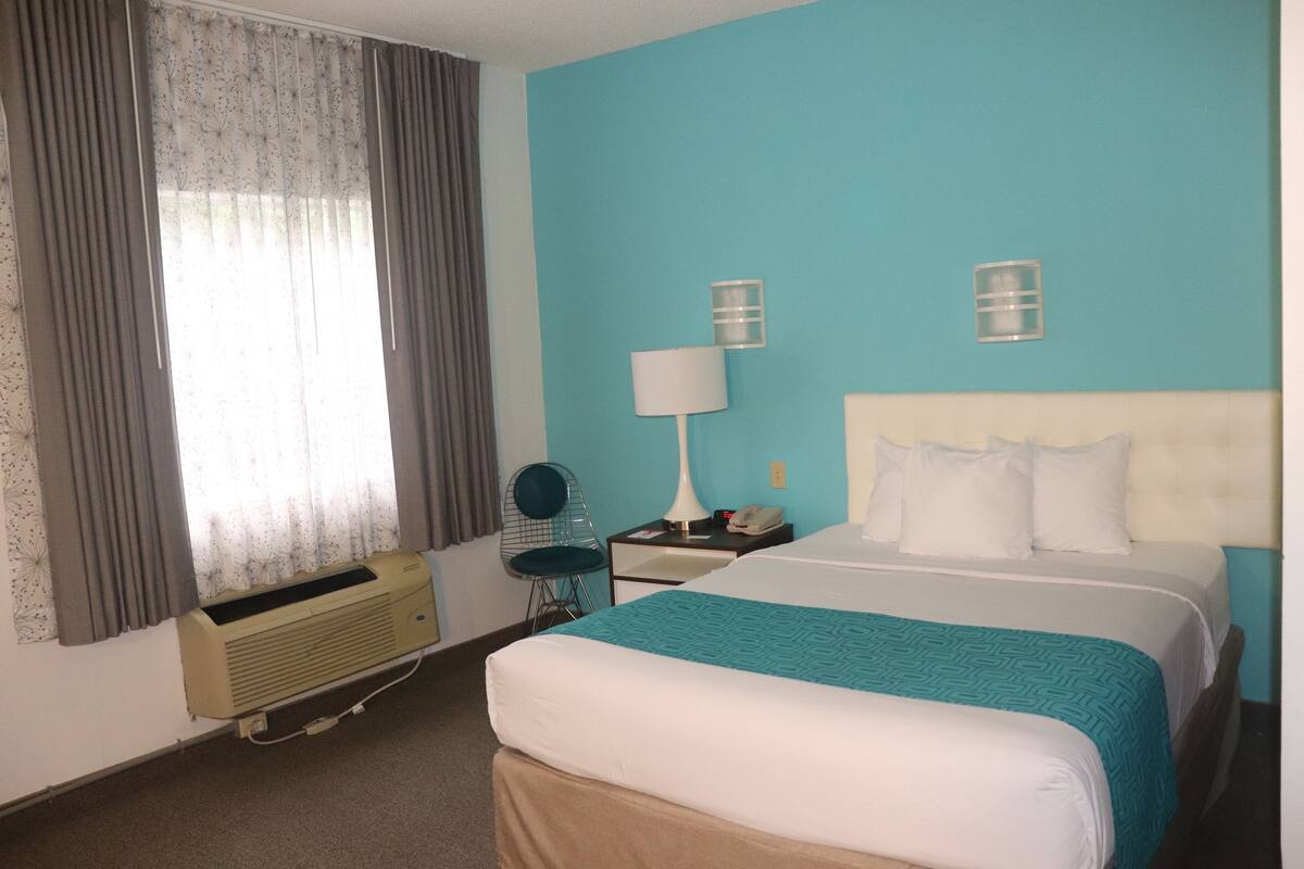 Quarto de hotel com cama, escrivaninha, cadeira, luminária e ar condicionado.
