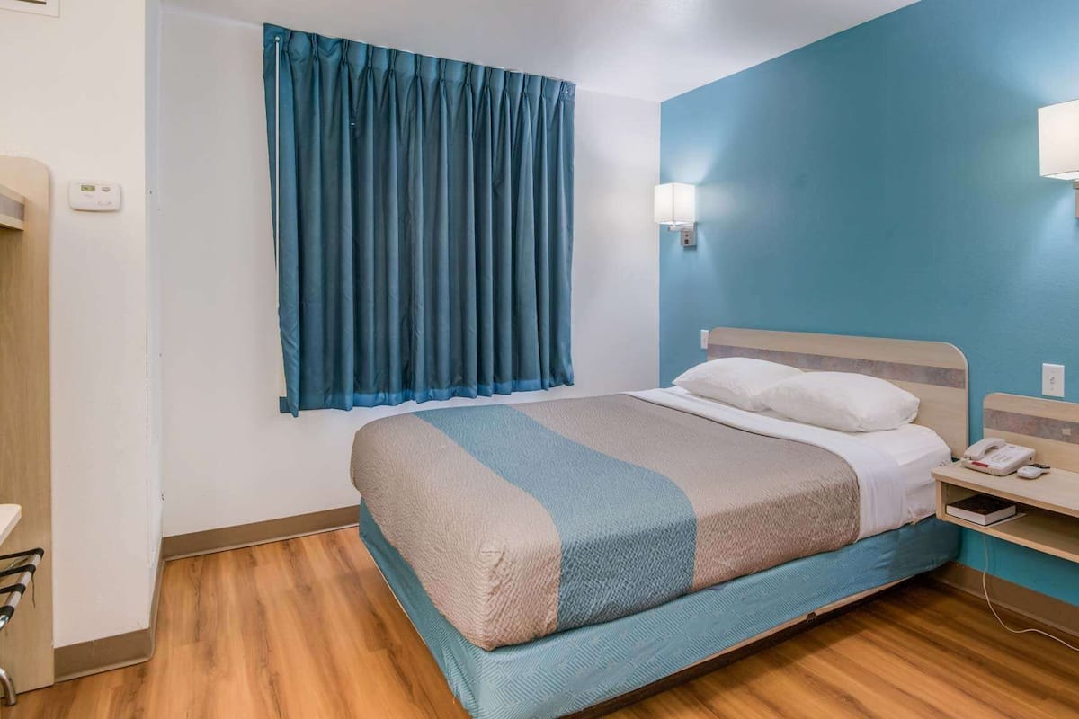 A hotel room with a bed, a bedside table with a lamp, a window with curtains, and a thermostat on the wall.