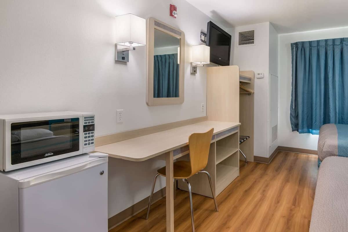 A compact hotel room with a bed, a desk, a chair, a microwave, a TV, and a window with curtains.