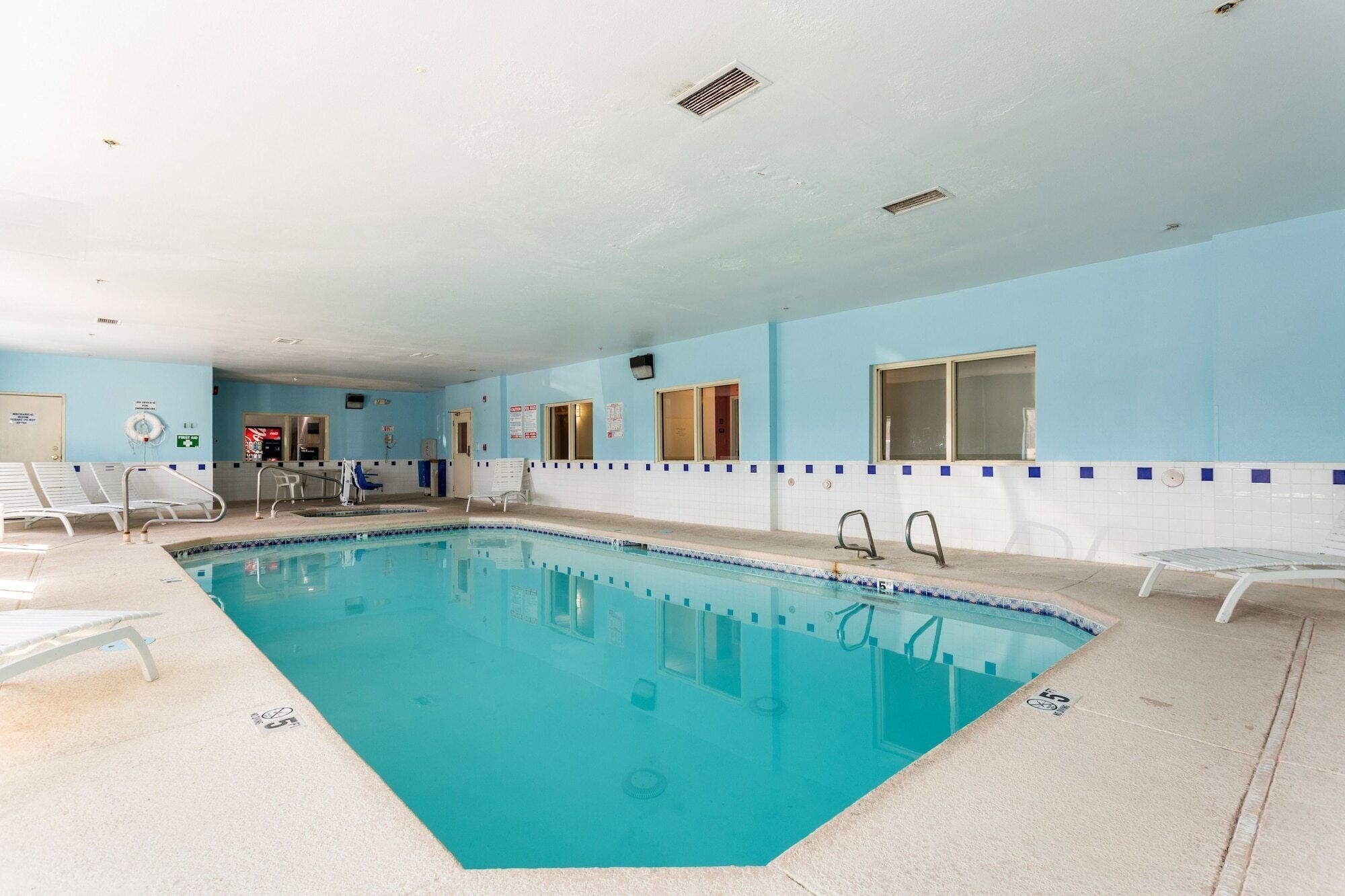 An indoor swimming pool with clear water, surrounded by lounge chairs and a tiled wall.