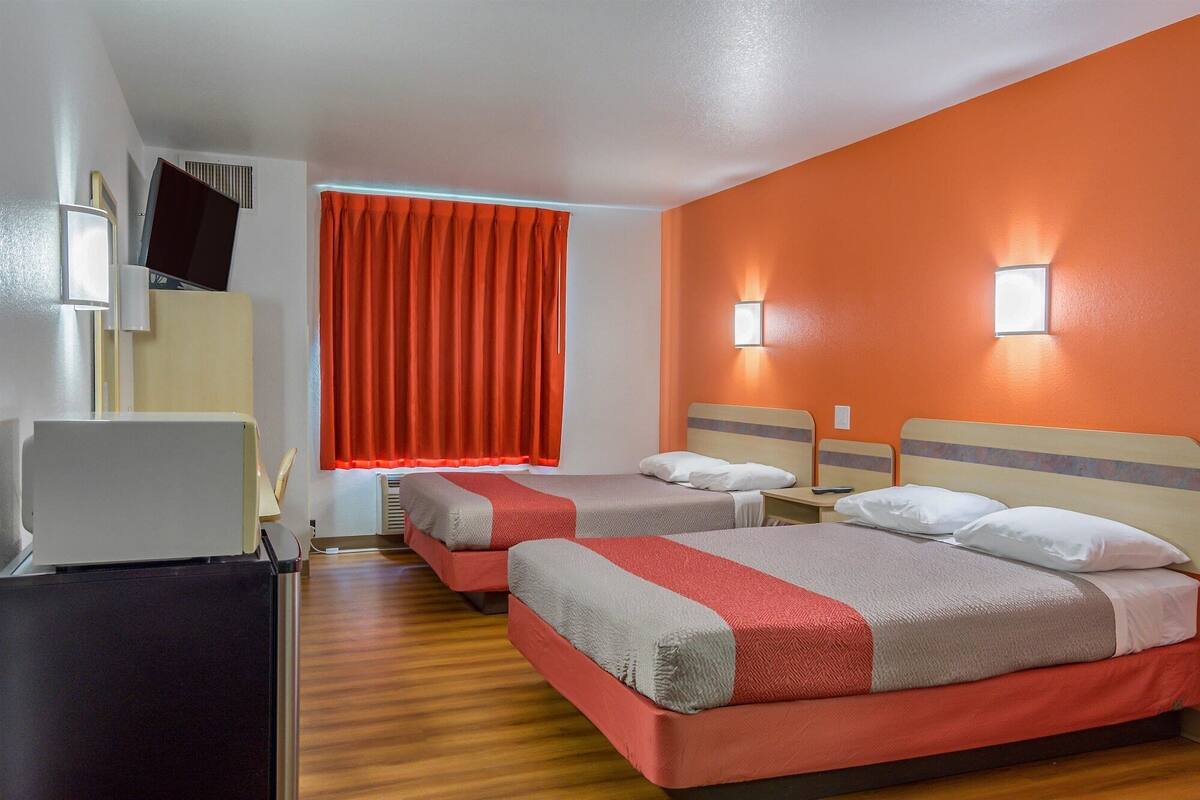 A hotel room with two beds, a desk, a chair, and a window with orange curtains.