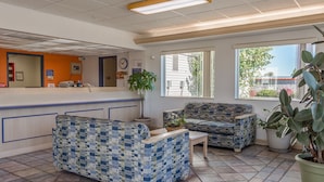 A reception area with a check-in counter, two patterned sofas, a small table, and a large potted plant. - Motel 6 Redmond, OR (Redmond)