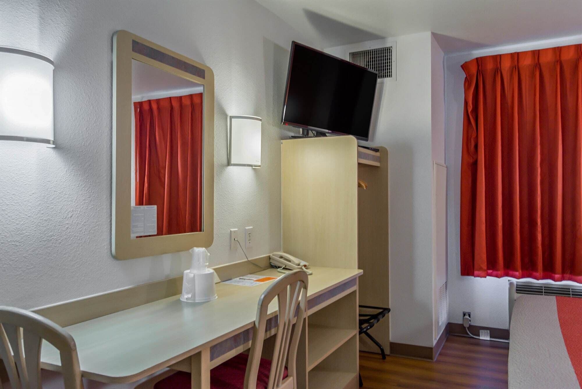 A hotel room with a desk, chair, mirror, television, wardrobe, and red curtains.