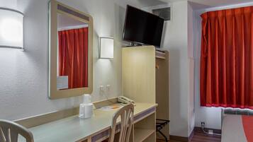 A hotel room with a desk, chair, mirror, television, wardrobe, and red curtains.
