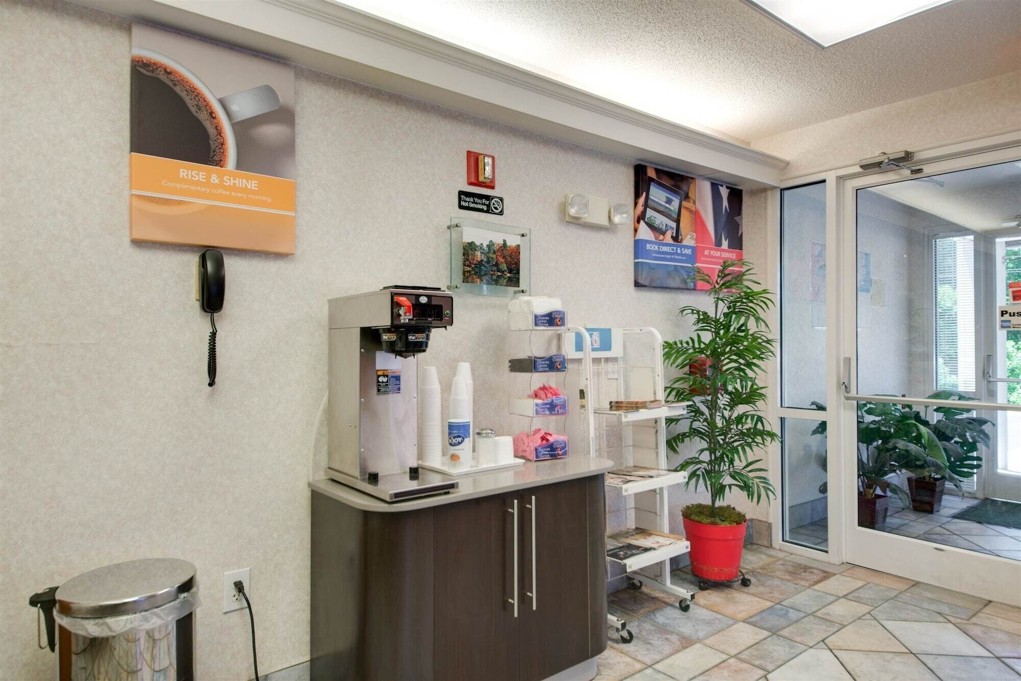 A reception area with a coffee machine, a trash bin, a plant, and a sign for 'RISE & SHINE' coffee.