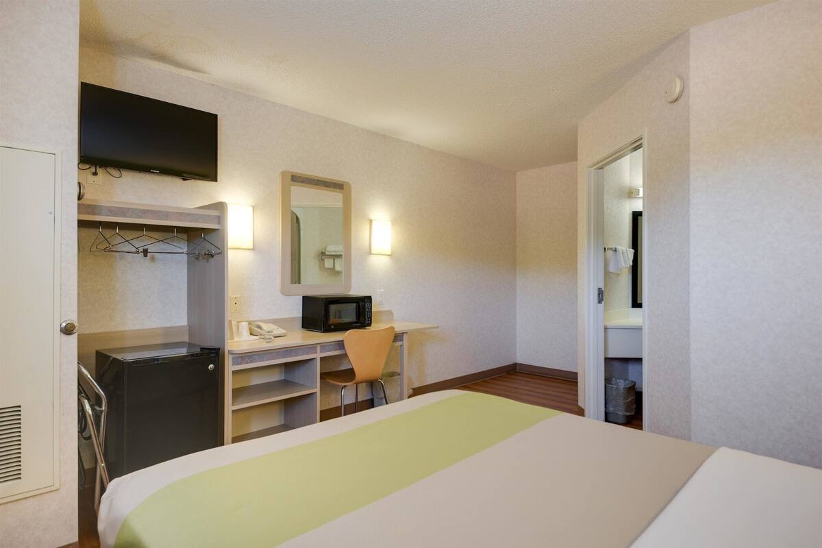 A hotel room with a bed, a desk with a chair, a microwave, a telephone, and a mirror.