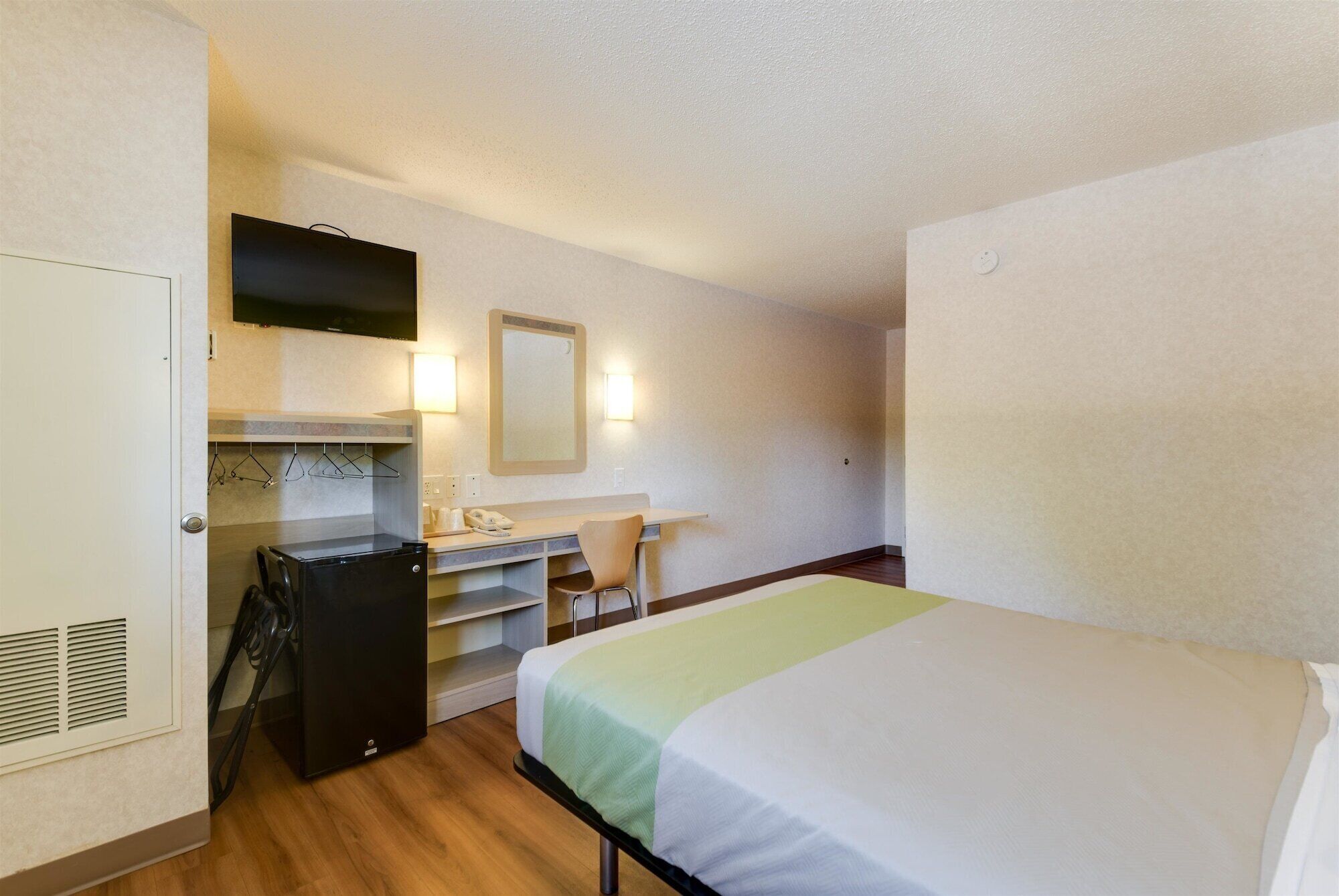 A compact hotel room with a bed, a desk with a chair, a wardrobe, a television, and a mirror.