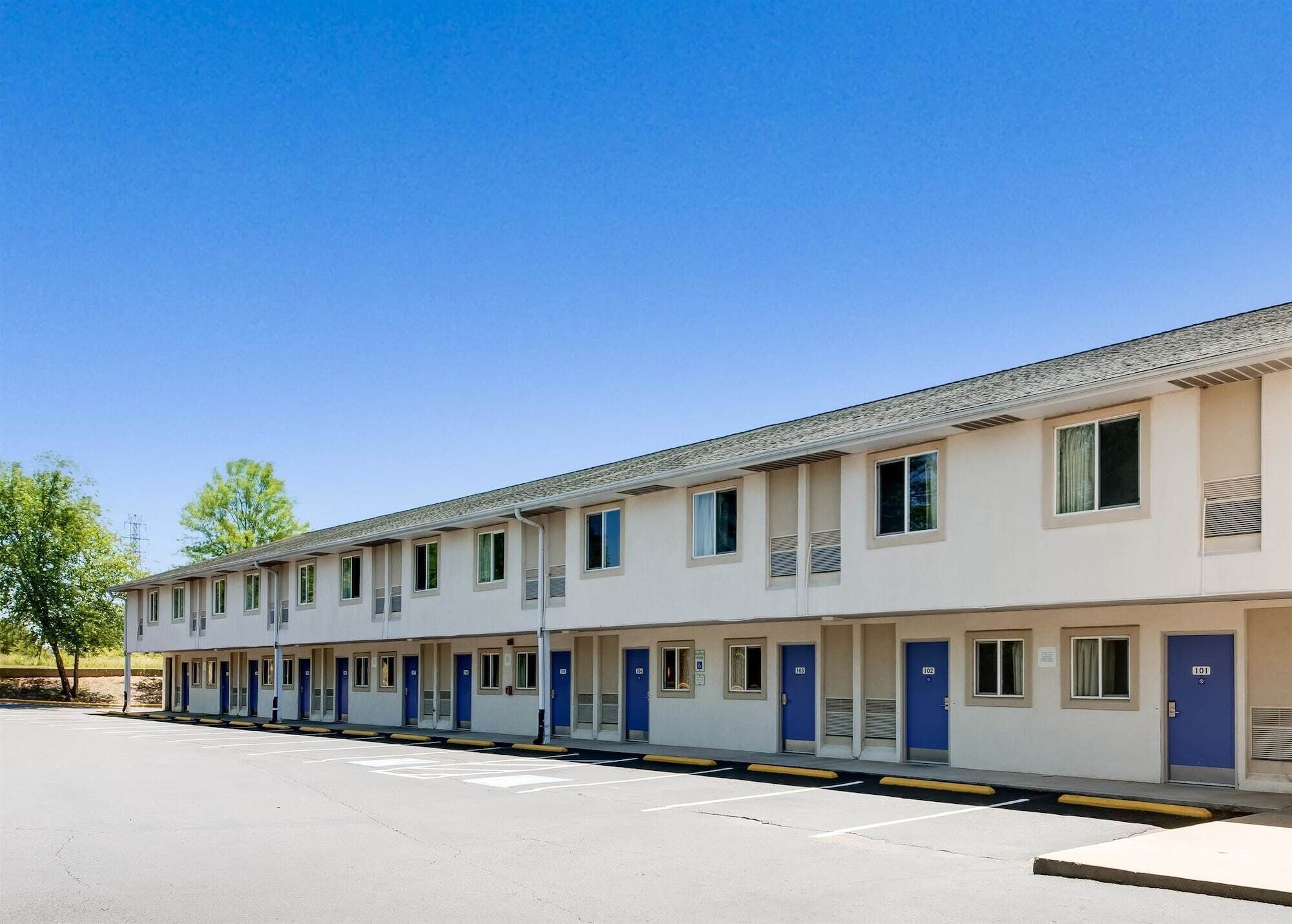 A row of single-story motel rooms with individual doorways and small windows, a parking lot in front, and a clear blue sky.
