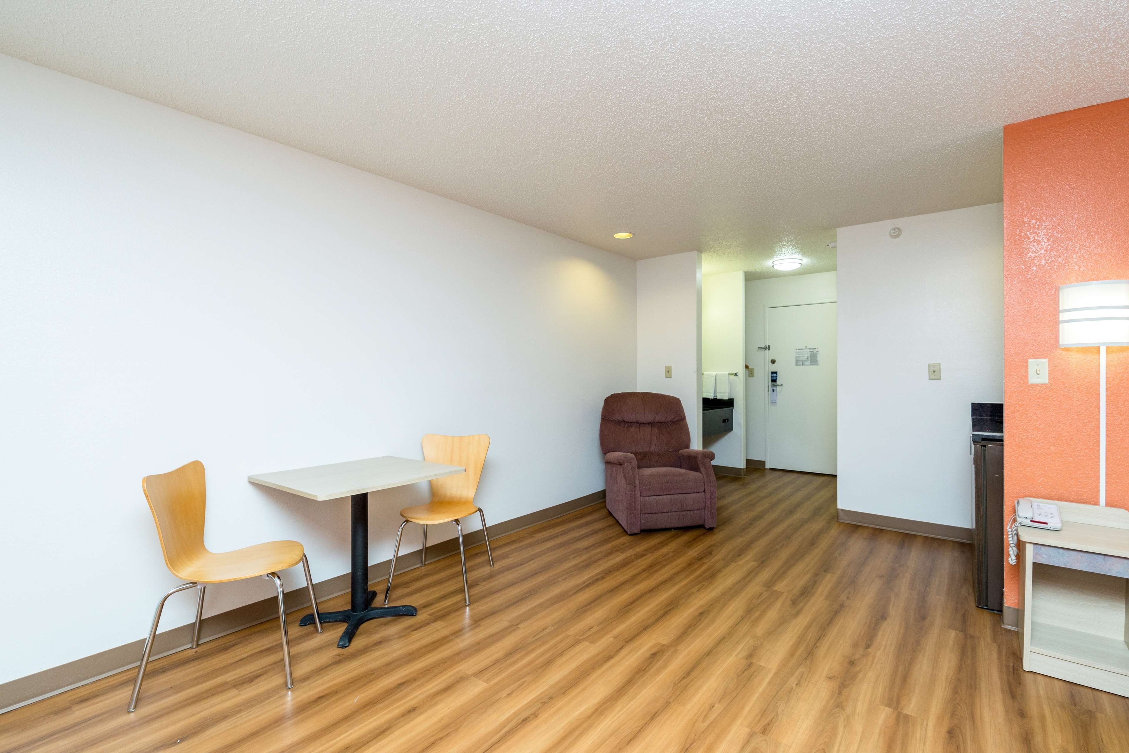 Una habitación con piso de madera, una mesa blanca, dos sillas, un sillón morado y un escritorio pequeño con un teléfono.