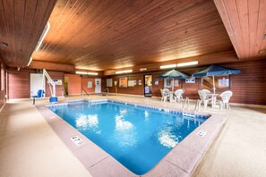 An indoor swimming pool with a wooden ceiling, clear pool area, and several tables and chairs around the pool.