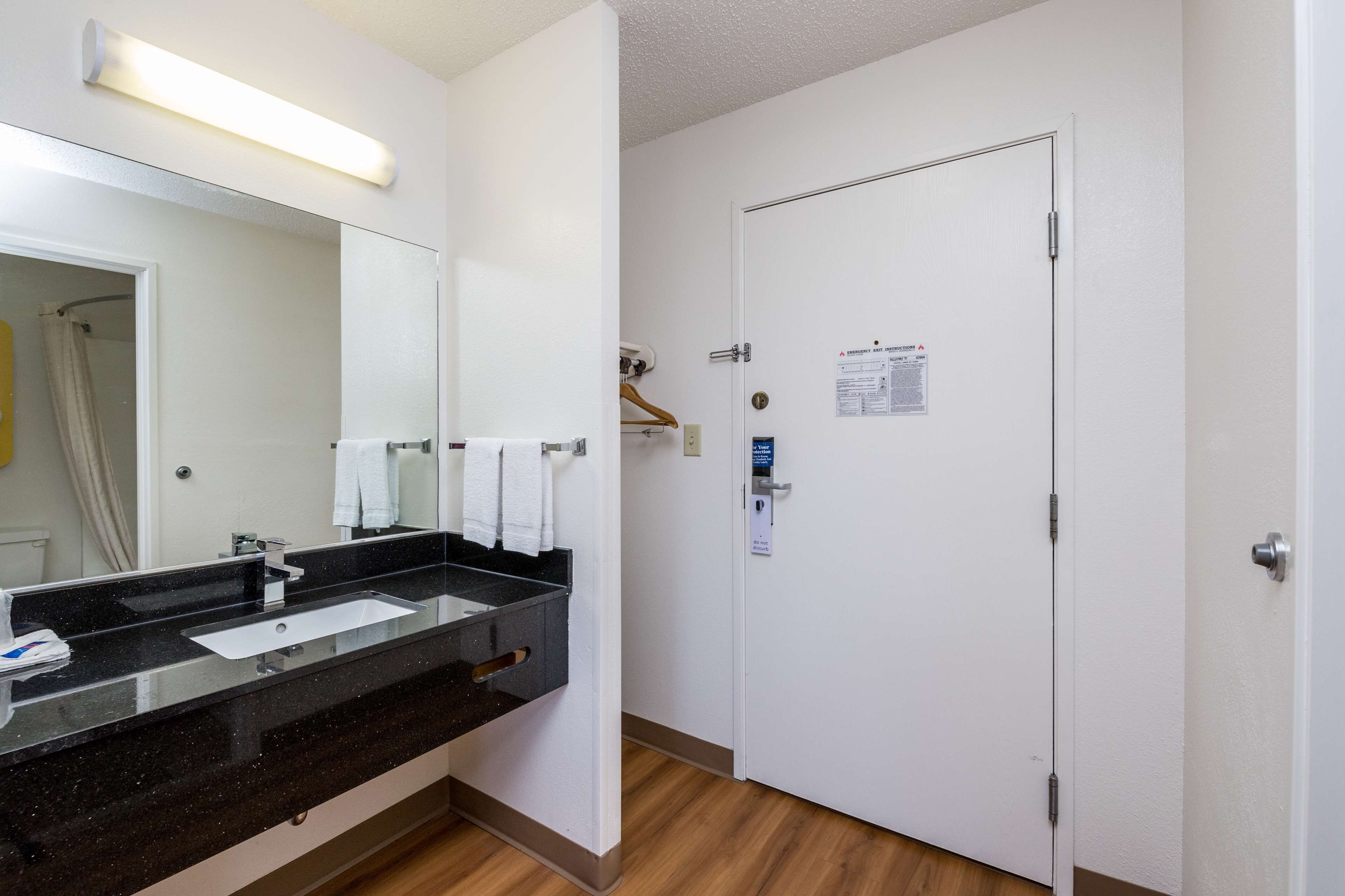 A hotel bathroom with a black countertop sink, a mirror, and a white door with a notice on it.