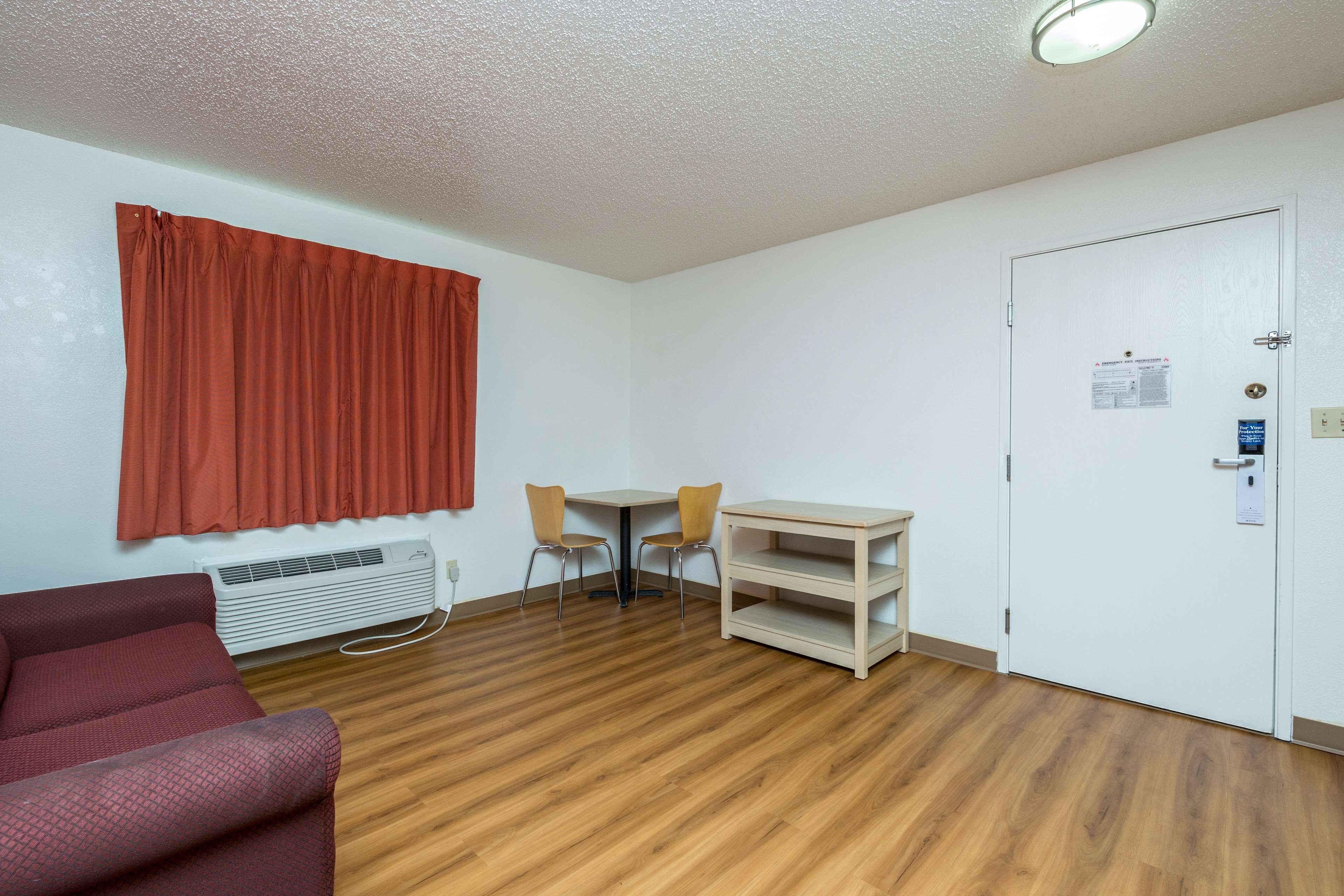 Una habitación con piso de madera, una cortina roja, una puerta blanca, una pequeña mesa con dos sillas y un sofá rojo.