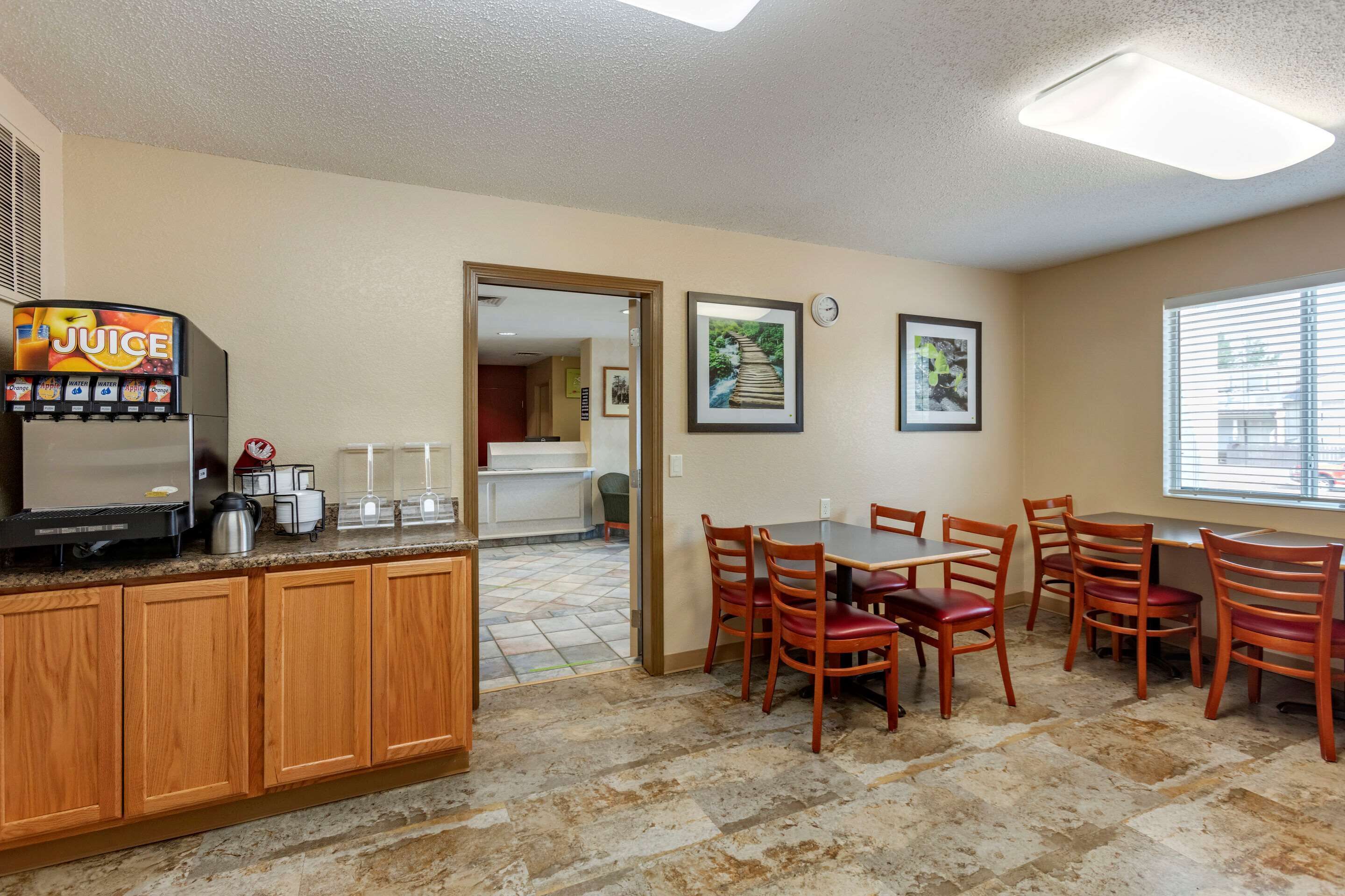 A hotel kitchen with a juice dispenser, a coffee maker, and a tiled floor. There is a dining area with wooden chairs and a table.