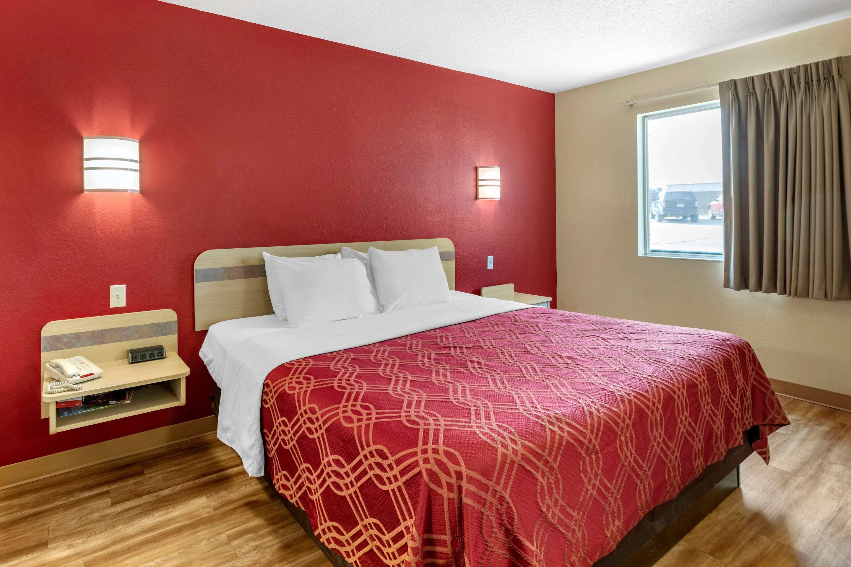 A hotel room with a red wall, a wooden floor, a bed with white bedding, a bedside table with a phone and books, and a window with curtains.