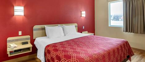A hotel room with a red wall, a wooden floor, a bed with white bedding, a bedside table with a phone and books, and a window with curtains.