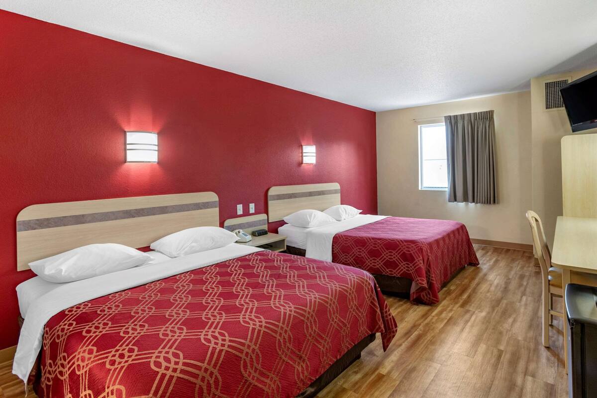 A hotel room with two beds, each with a red patterned bedspread, a wooden headboard, and a nightstand with a lamp.