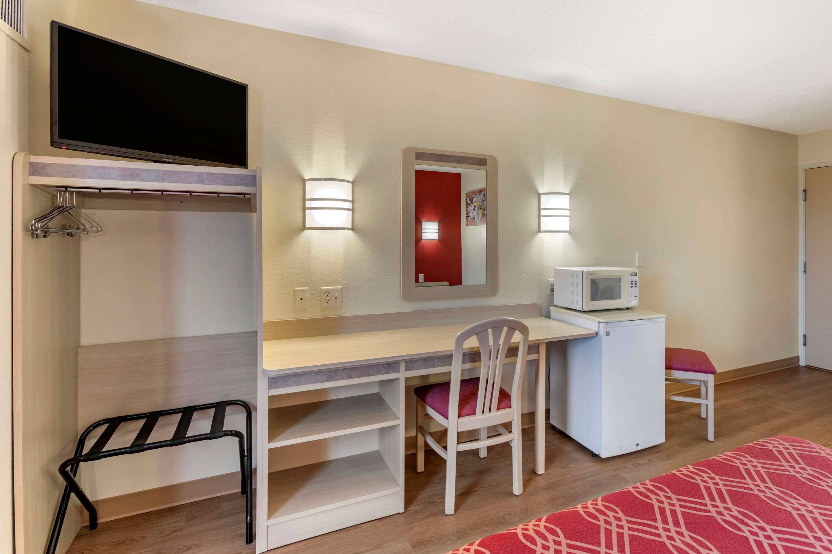A compact room with a bed, a desk with a chair, a TV mounted above the desk, a microwave, and a small refrigerator.