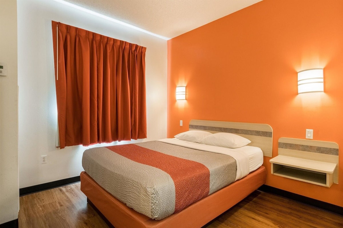 A hotel room with a bed, a nightstand, a wall-mounted light, and a window with curtains.