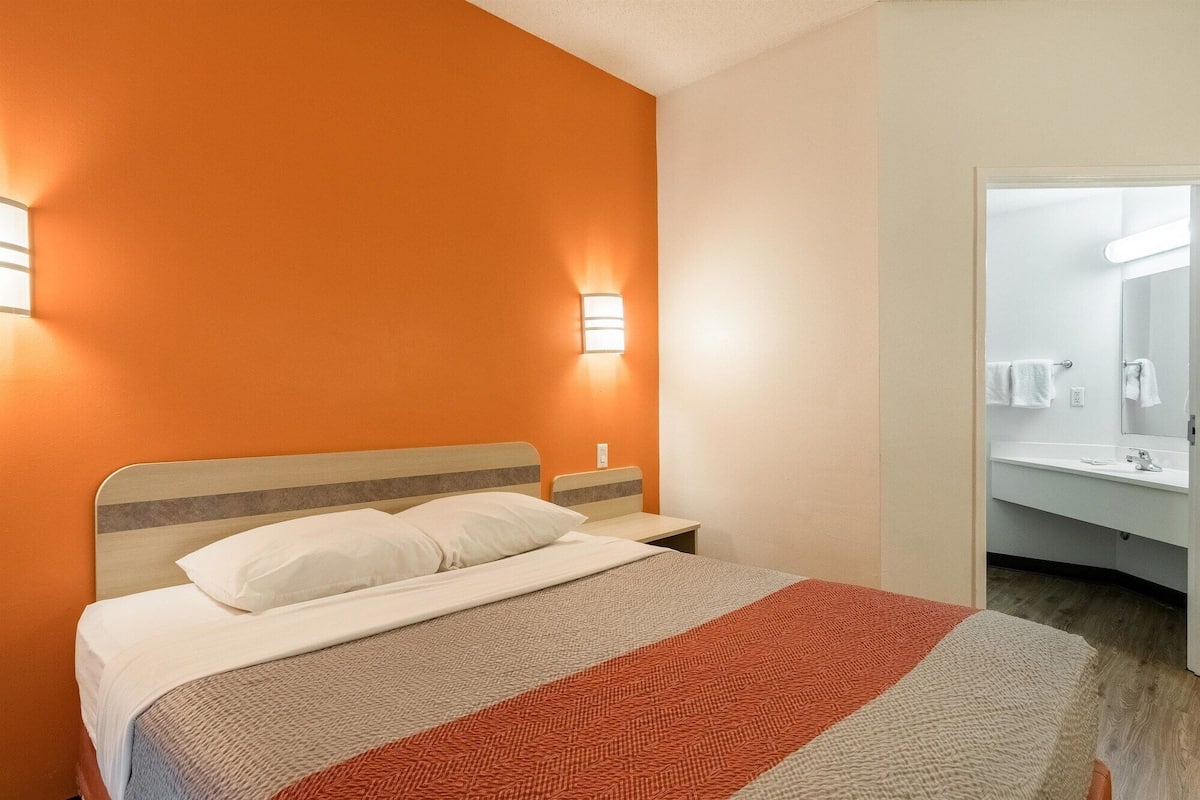A hotel room with a bed, a nightstand, a wall-mounted light, and a bathroom visible through an open door.