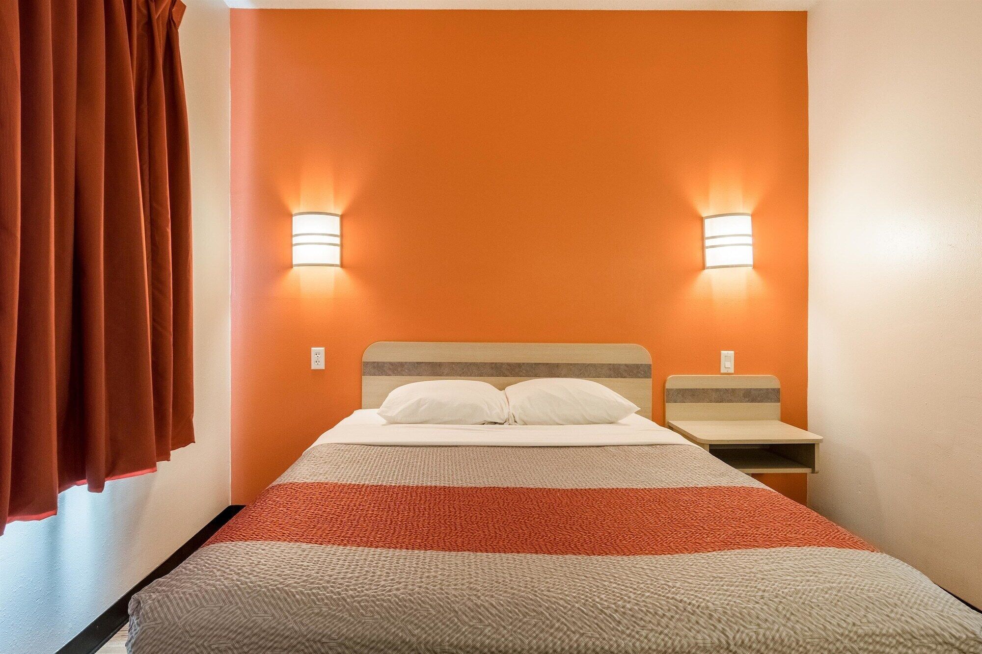 Habitación de hotel con pared de acento naranja, una cama con manta a rayas, dos mesitas de noche y lámparas fijadas a la pared.