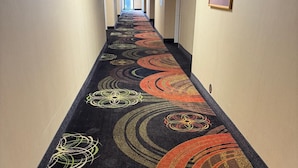 Un pasillo de hotel con alfombra estampada, un cuadro en la pared y una puerta al final.