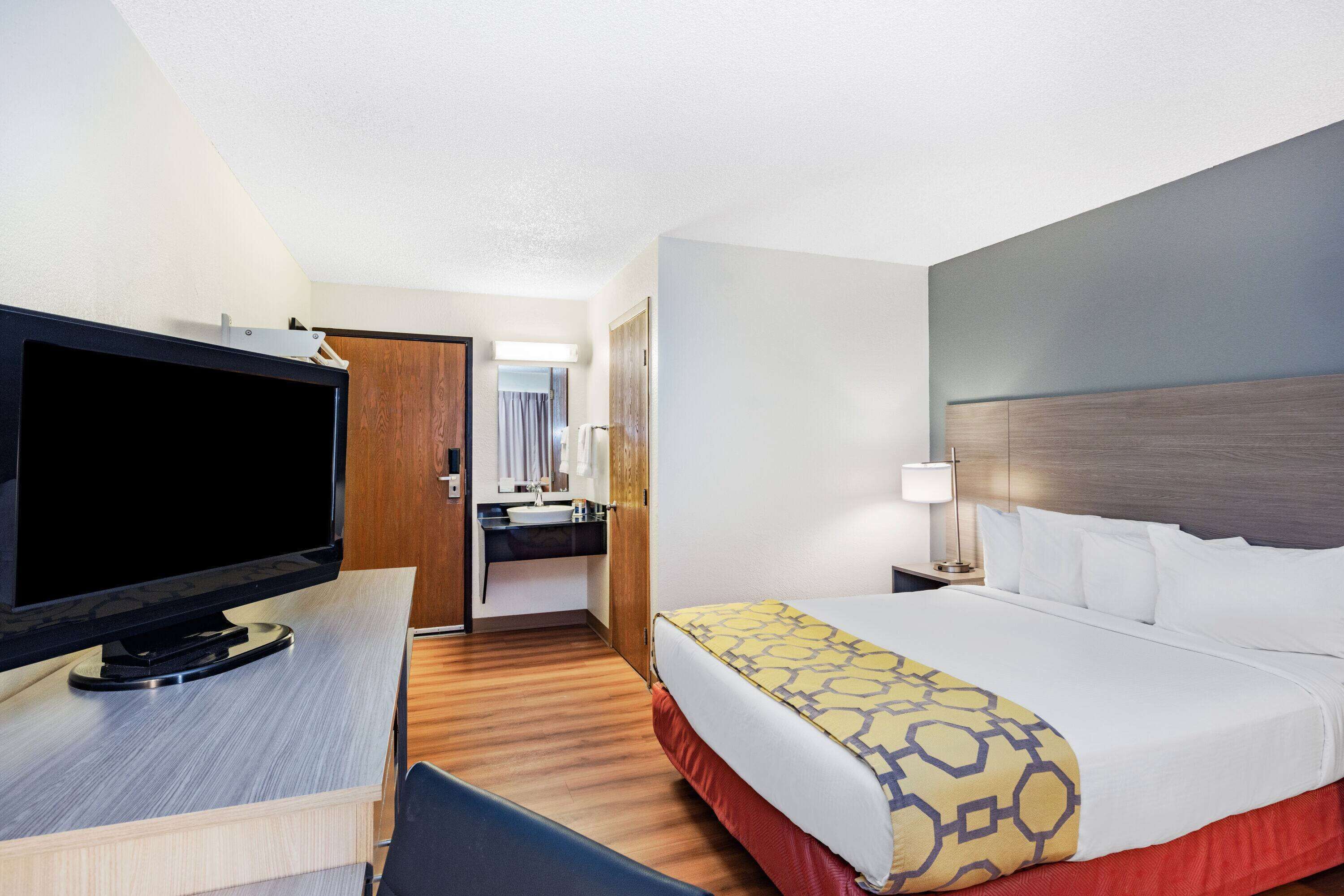 A hotel room with a bed, a desk with a flat-screen TV, a bathroom with a mirror and sink, and a door leading to another room.