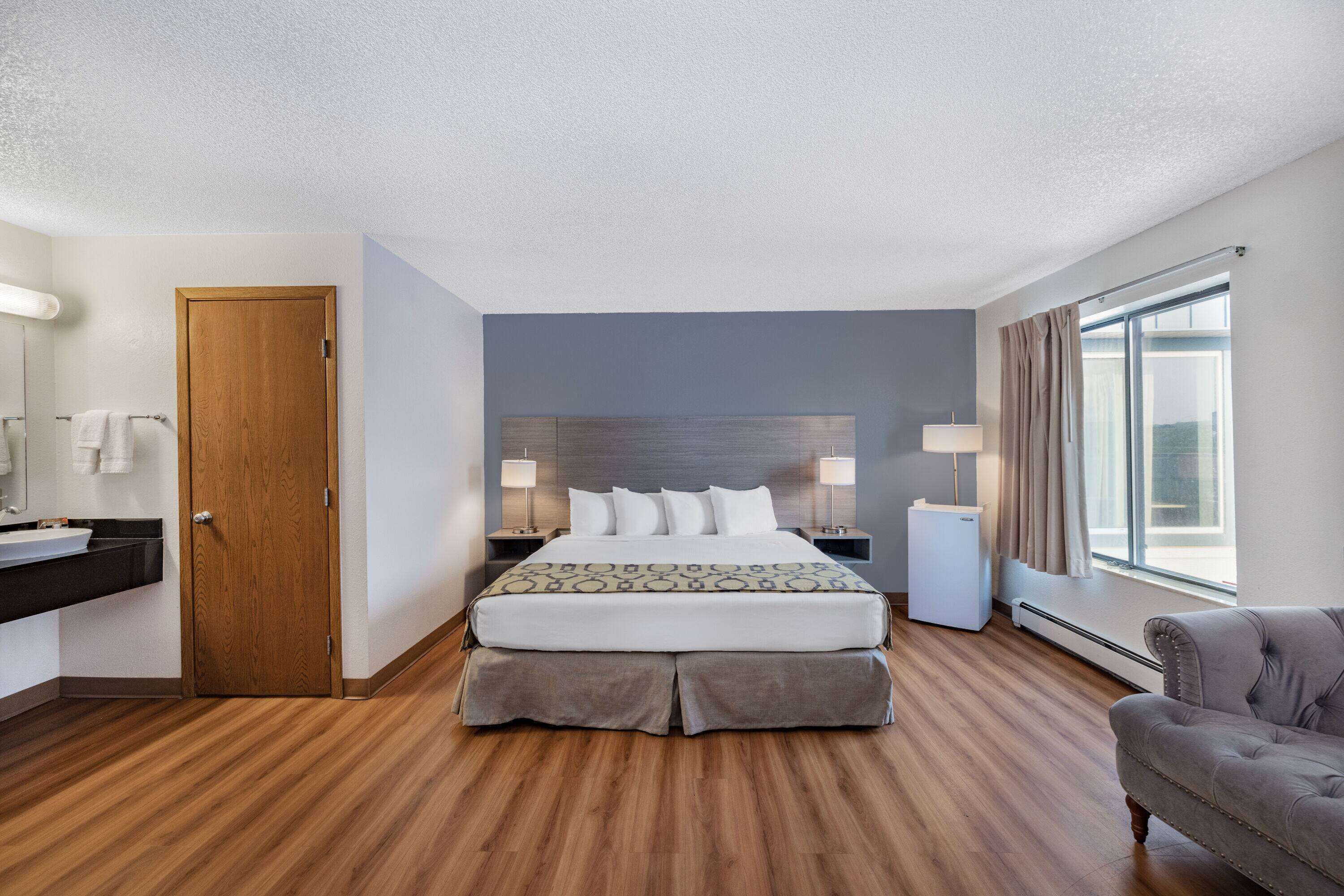 A hotel room with a wooden floor, a bed with white bedding, a grey armchair, a bedside table with a lamp, a small white cabinet, and a window with curtains.