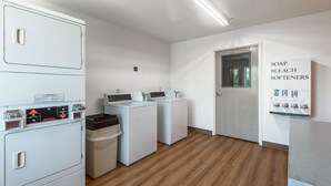A laundry room with two washing machines, a dryer, and a wall-mounted soap and bleach softener dispenser. - Motel 6 Junction City, KS (Junction City)