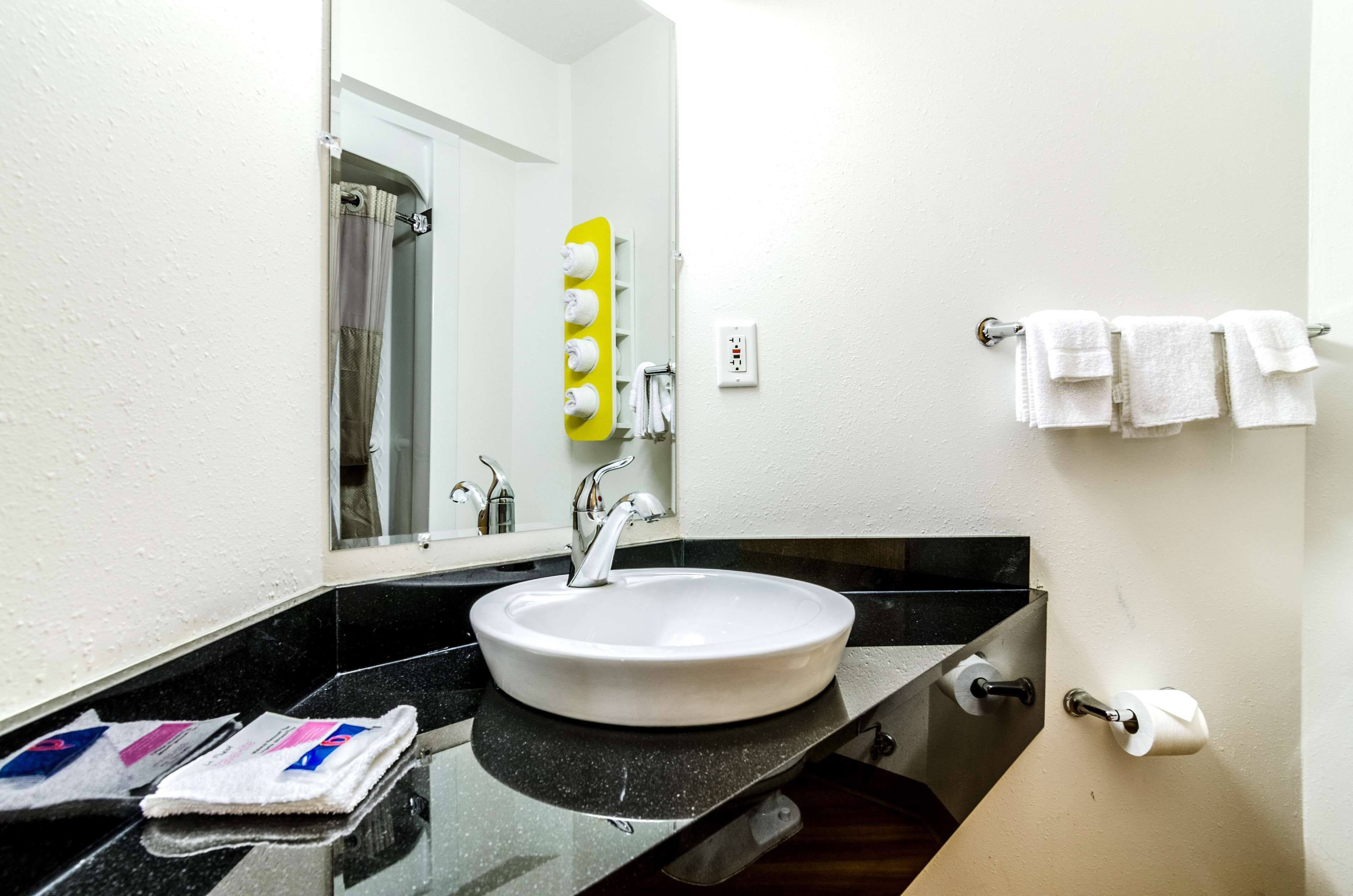 Une salle de bain avec un lavabo blanc, un plan de toilette noir et un miroir. On peut voir des serviettes accrochées au mur et une douche à l’arrière-plan.