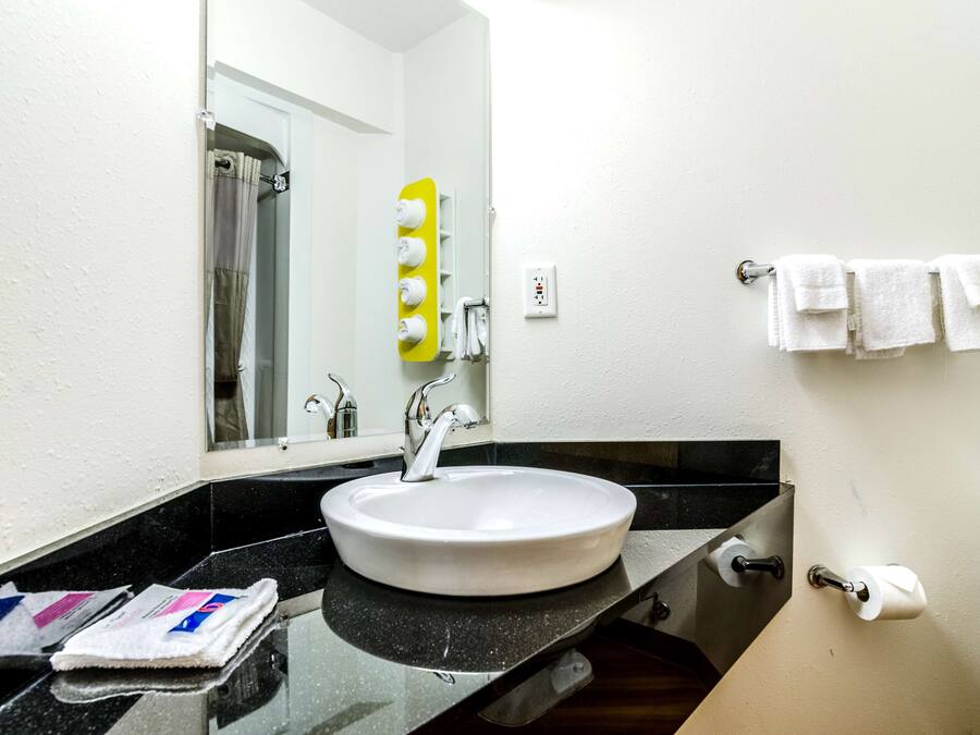 A bathroom with a white sink, black countertop, and a mirror. There are towels hanging on the wall and a shower visible in the background.