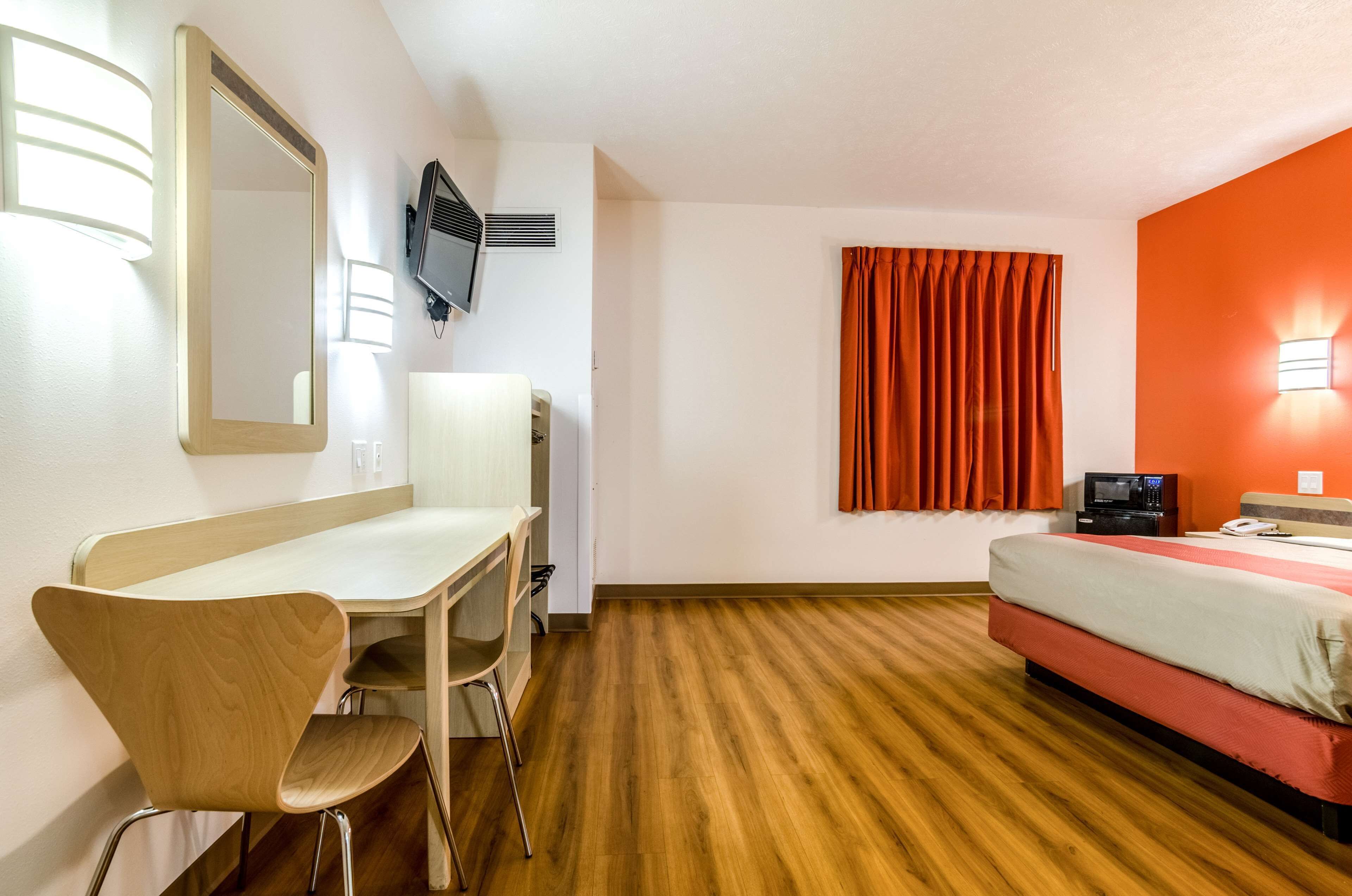 Ett hotellrum med trägolv, ett skrivbord med en stol, en säng, en tv och ett fönster med orangea gardiner.
