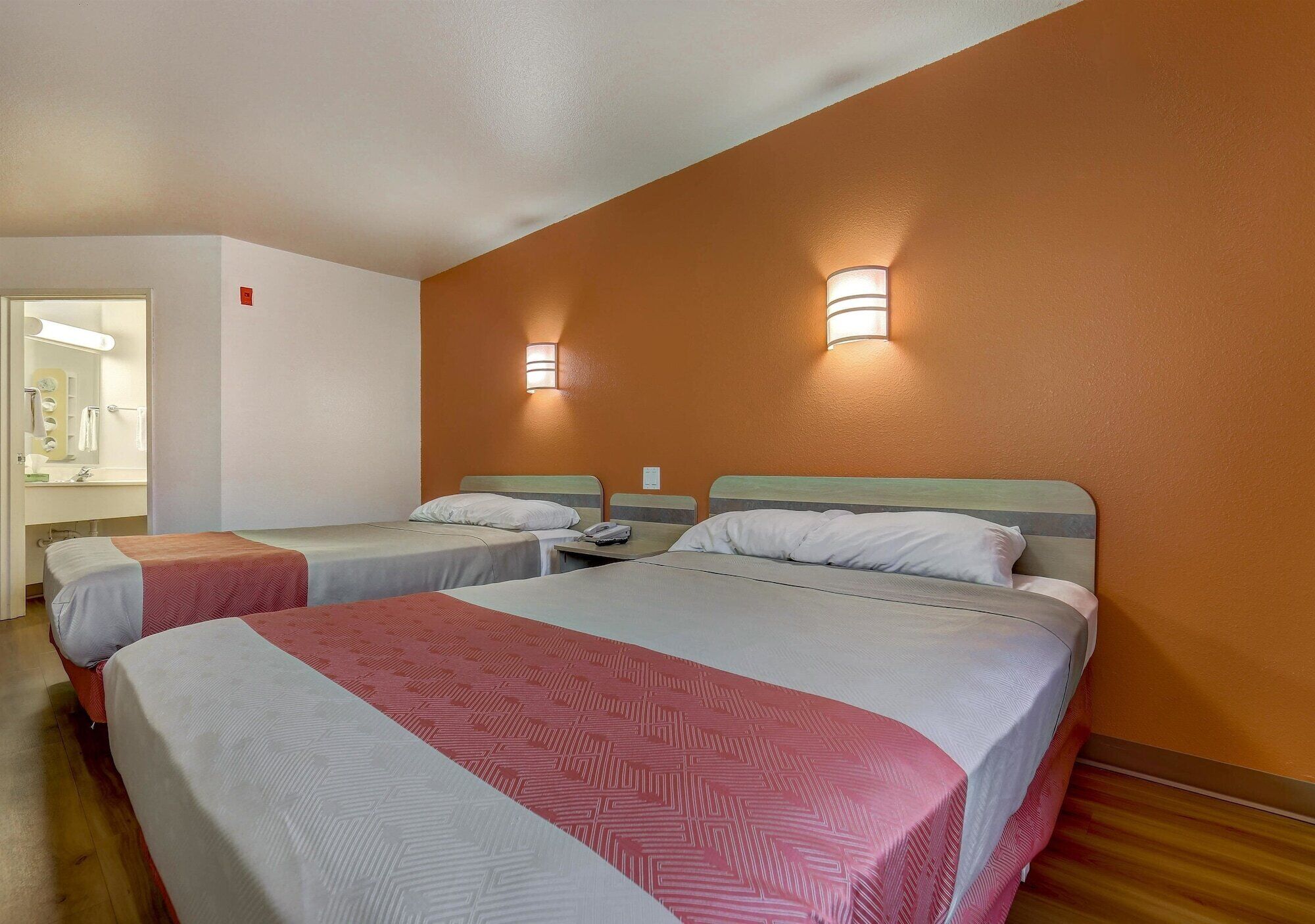 Quarto de hotel com duas camas, banheiro visível por uma porta aberta e luminárias fixadas na parede.