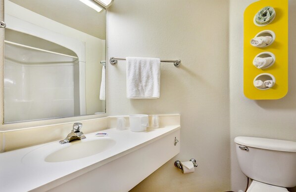 Un baño con lavamanos blanco, espejo y retrete. Hay un perchero con una toalla blanca y un dispensador de toallas amarillo, adosado a la pared, con varias toallas.