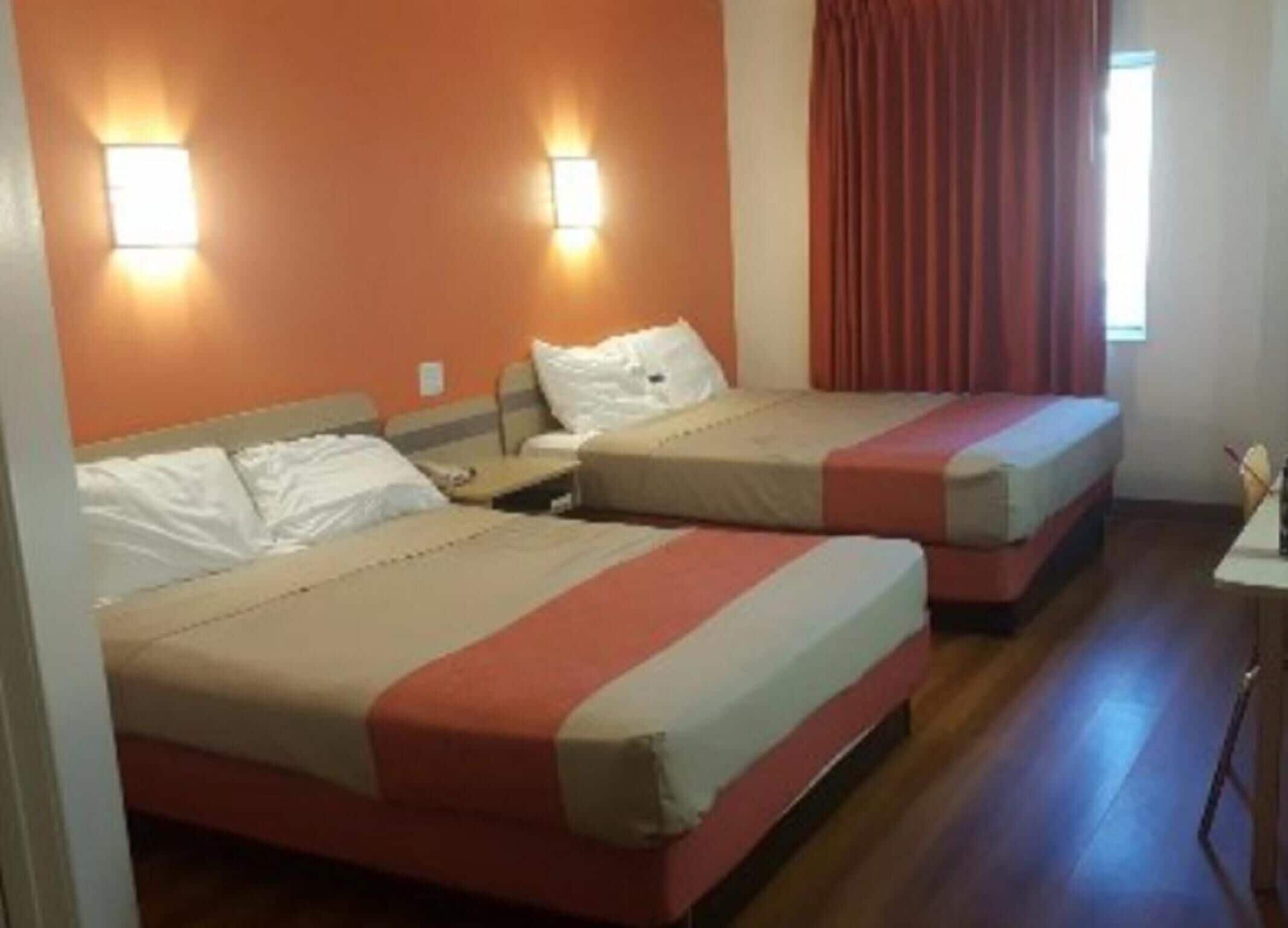 A hotel room with two beds, each with a headboard, a nightstand, and a lamp. The room has an orange wall and a window with red curtains.